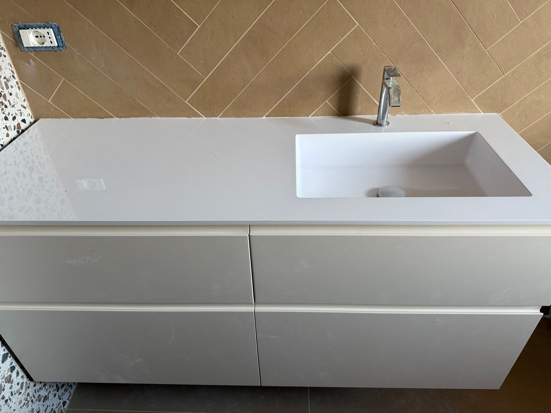 Un mobile da bagno bianco con lavabo e rubinetto integrati, addossato a una parete rivestita di piastrelle marroni con motivo a spina di pesce.
