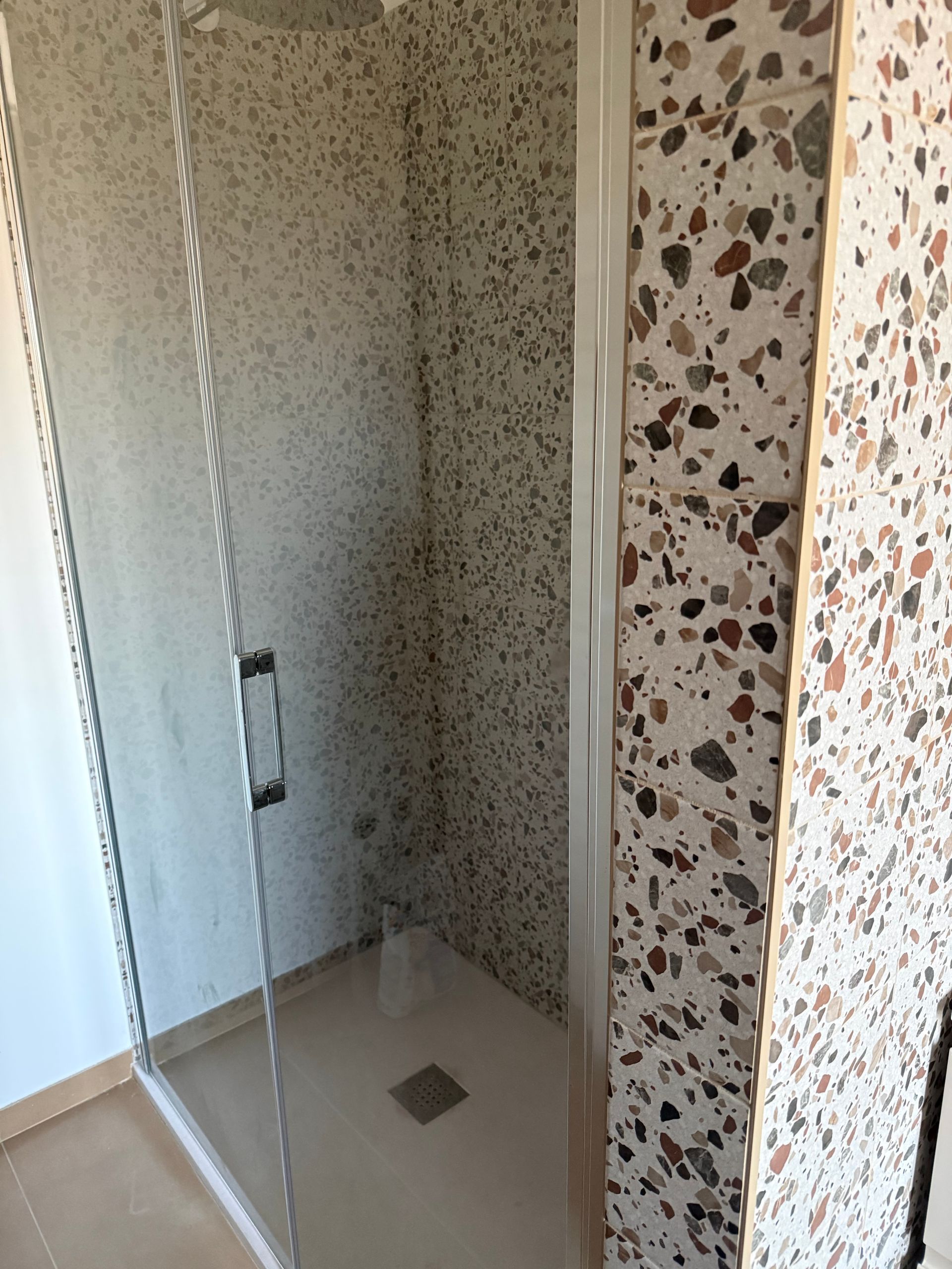 Box doccia con porta in vetro, caratterizzato da pareti rivestite con piastrelle in stile terrazzo di colore chiaro e frammenti di pietra multicolore.