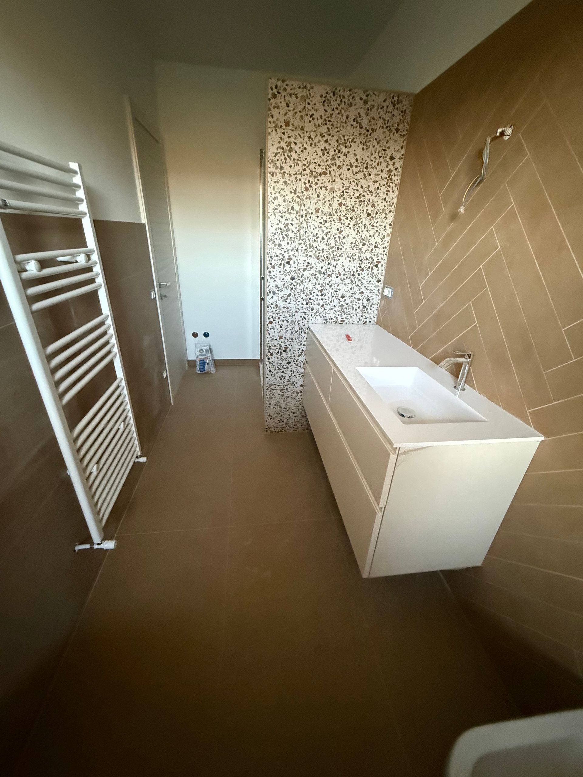 Un bagno moderno con piastrelle beige a spina di pesce, un mobile lavabo bianco, una parete decorativa in terrazzo e uno scaldasalviette bianco.