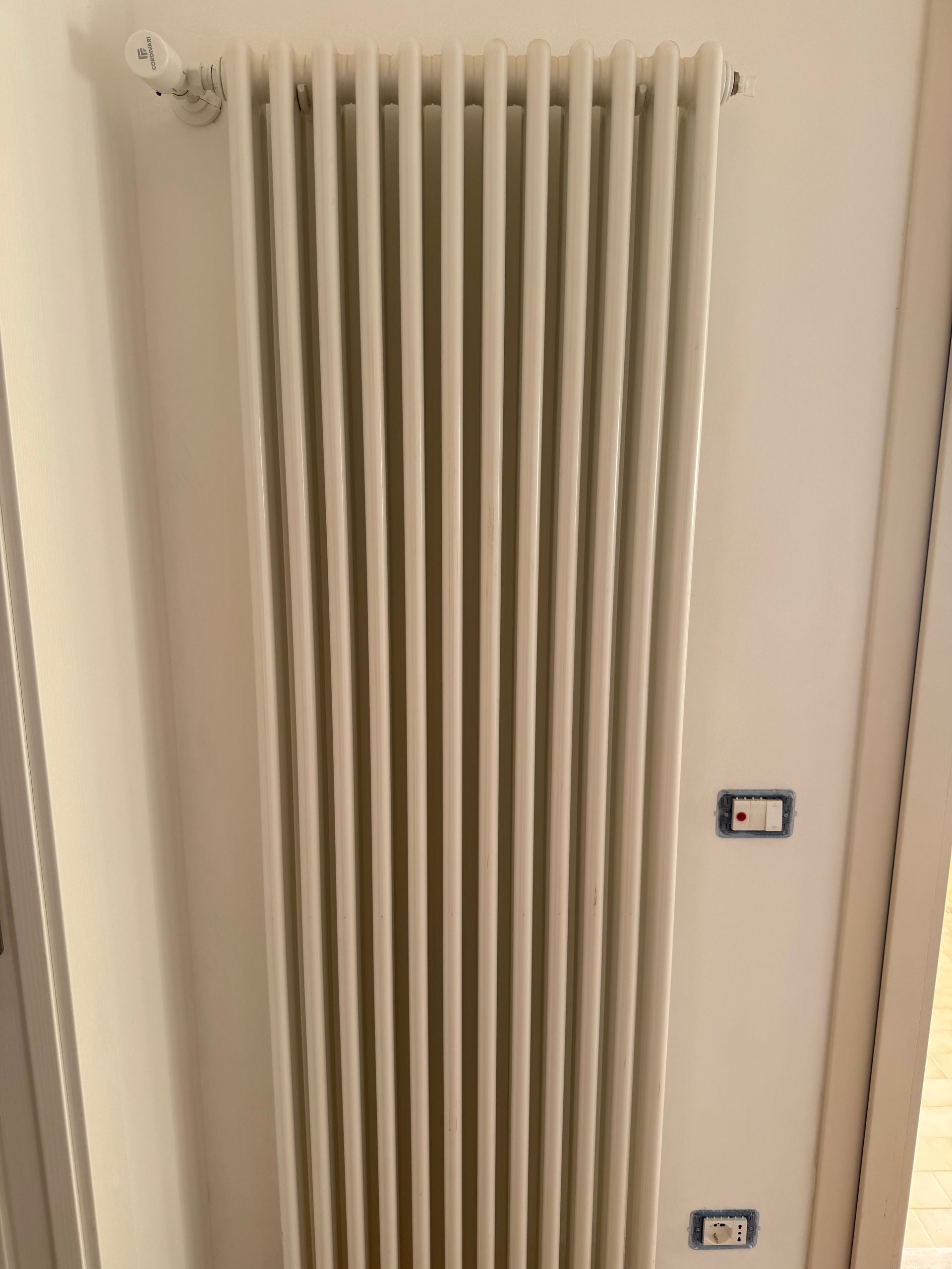 Un radiatore alto e verticale di colore bianco, montato su una parete bianca accanto a due prese elettriche.