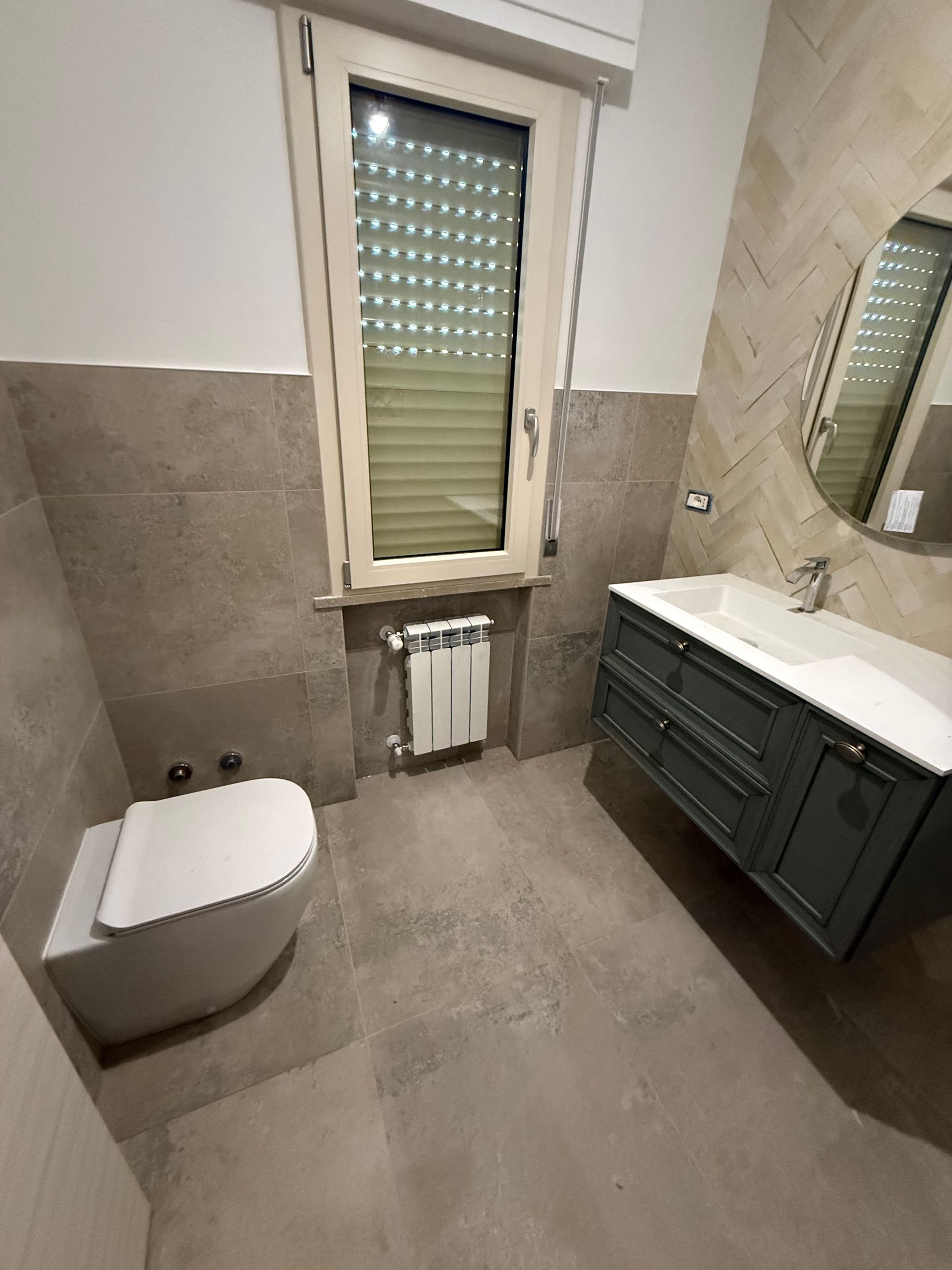 Bagno moderno con piastrelle grigio chiaro a pavimento e a parete, WC sospeso bianco, mobile lavabo grigio scuro e finestra.