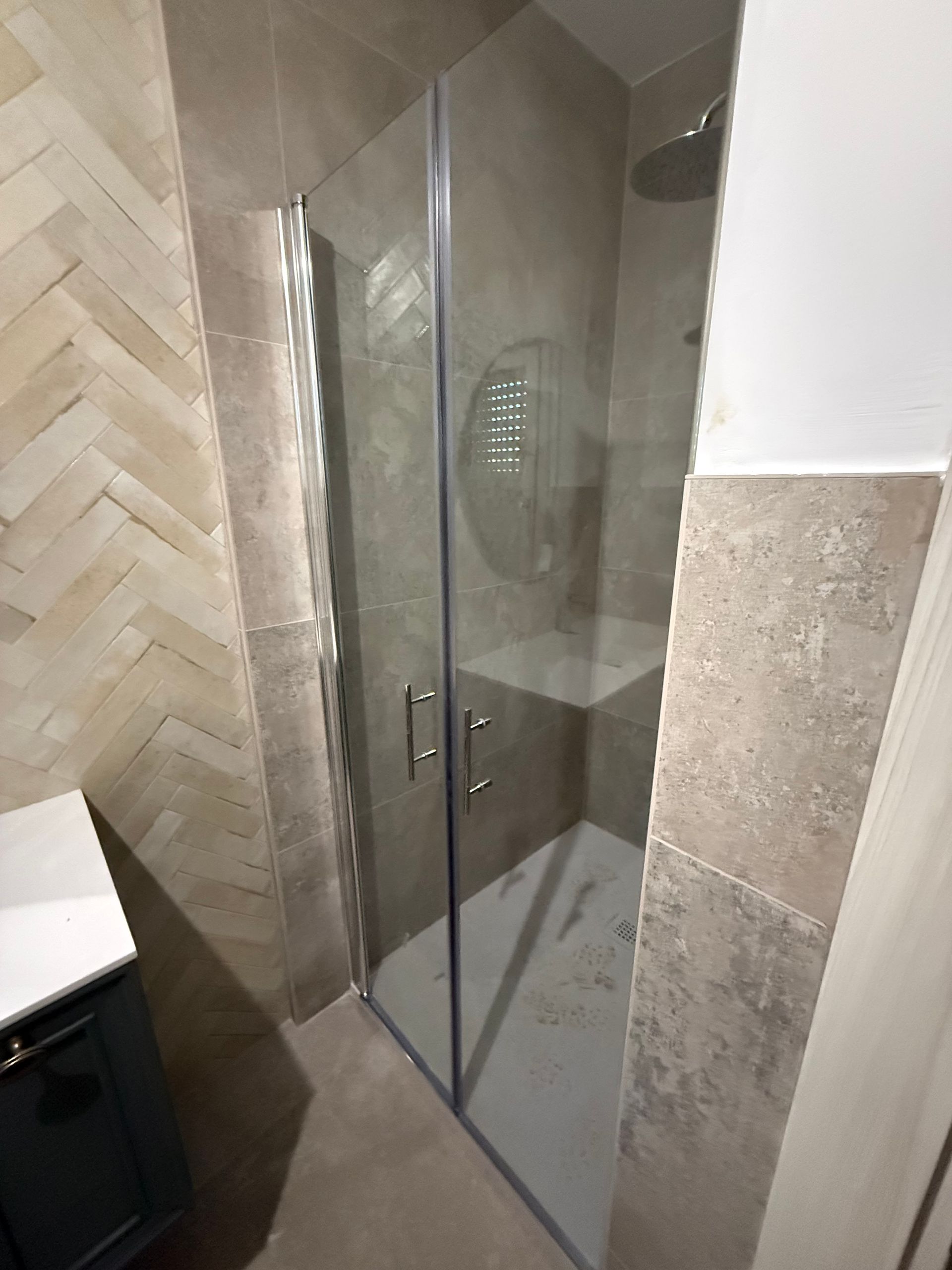 Una cabina doccia in vetro con piastrelle a spina di pesce beige sulle pareti e rivestimento in piastrelle di pietra dai toni neutri.