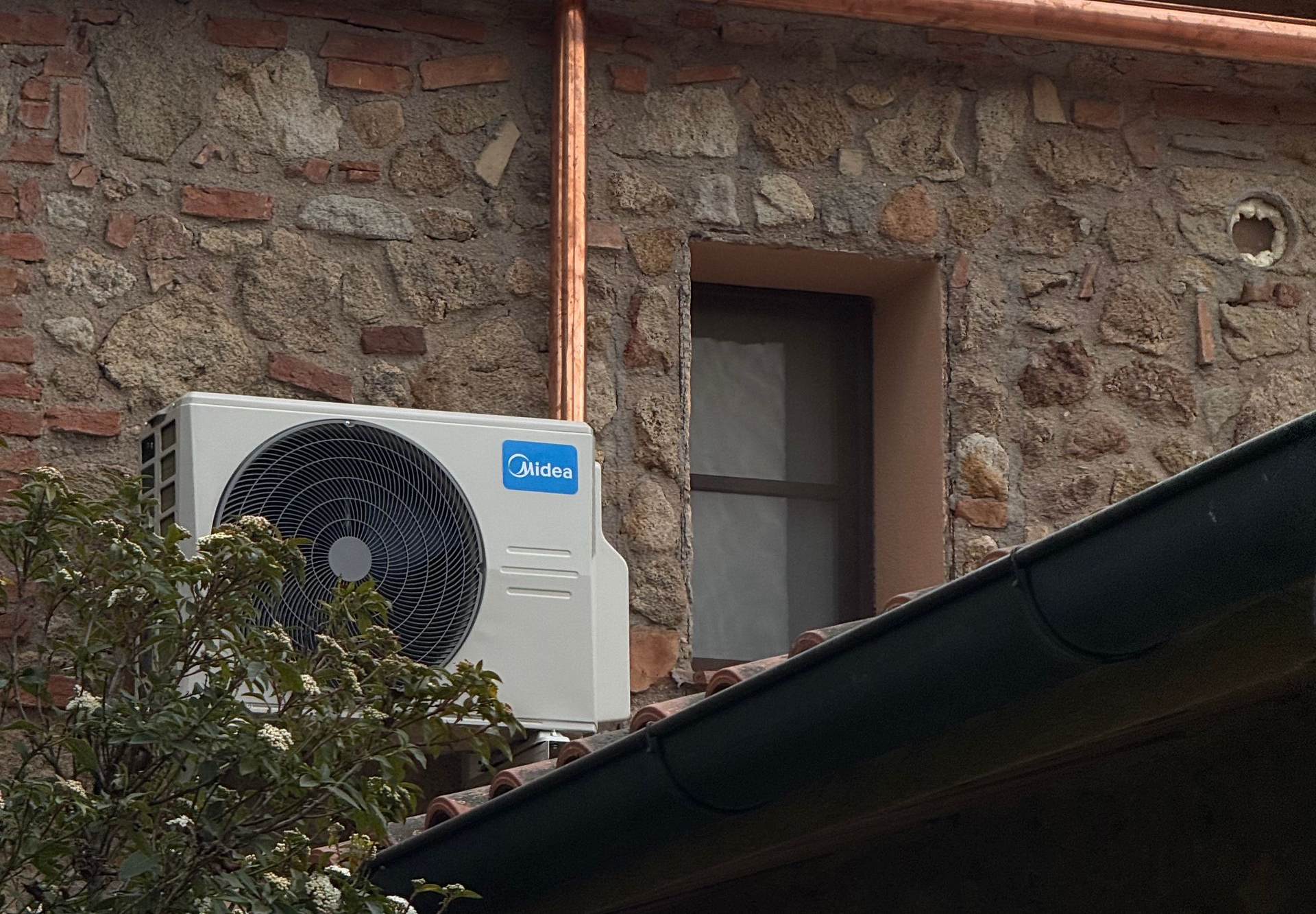 Un condizionatore d'aria bianco di marca Midea, montato su un muro esterno in pietra rustica accanto a una finestra incassata.