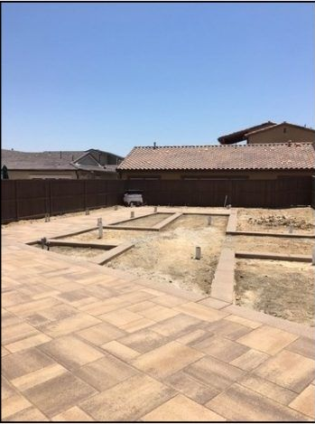 Backyard with paved patio, garden beds, and brown fence under a blue sky.