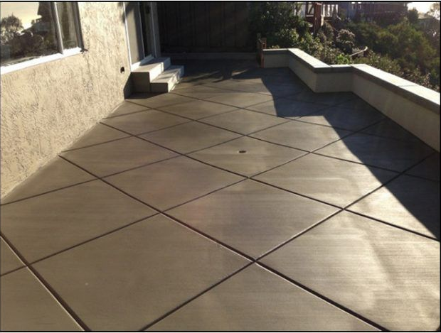 Concrete patio with diamond pattern, next to a stucco wall, with steps and a low retaining wall overlooking greenery.