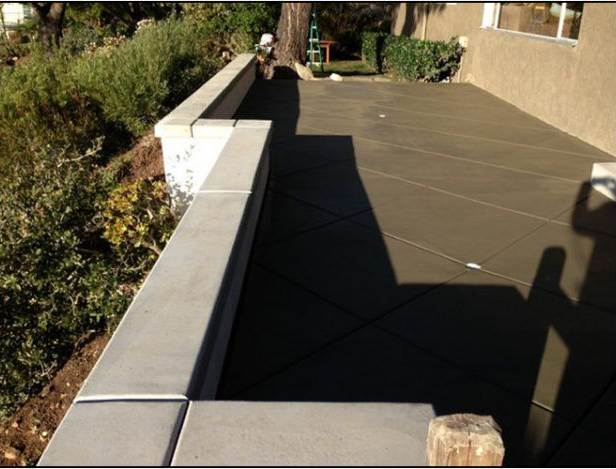 Newly poured concrete patio and retaining wall next to hillside, with a house in the background.