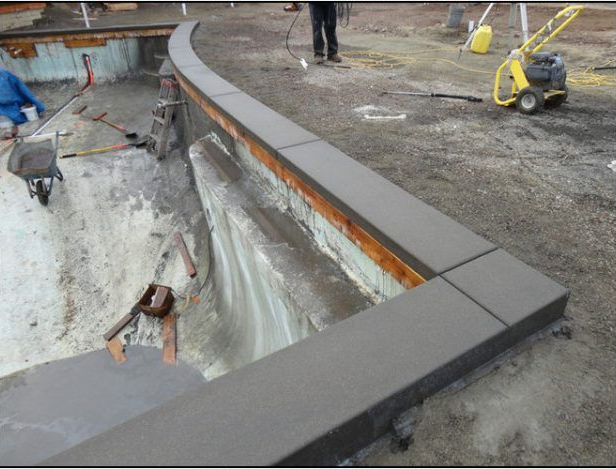 Construction site: curved concrete border being installed around a pool. Workers, tools, and a compactor visible.