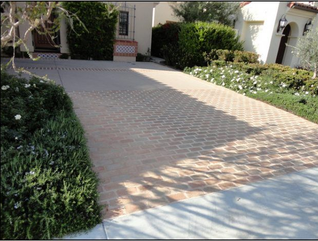 Driveway paved with brick-like pattern, bordered by bushes and flowers, leading to a house.