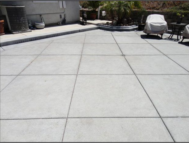 Large, light-colored concrete patio with grid lines, bordered by a building, plants, and covered outdoor furniture.