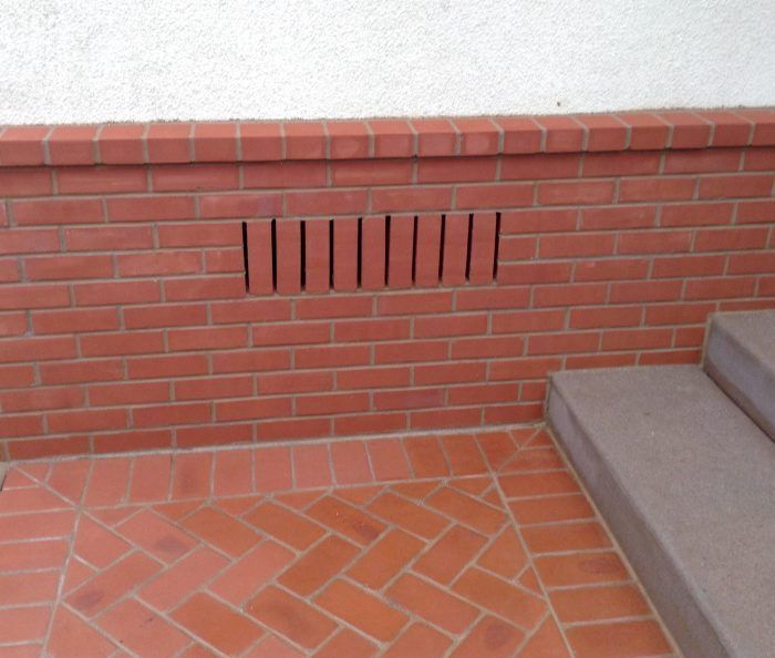 Red brick wall with ventilation slots, near tiled floor and concrete steps.
