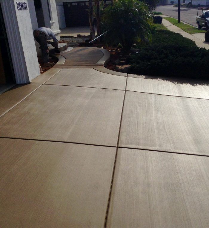 Man working on concrete driveway. Tan concrete with expansion joints. Green bush on the right.