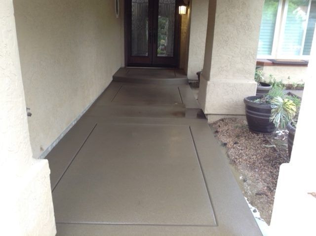 Concrete walkway leading to a front door, bordered by pillars, with potted plants on the side.
