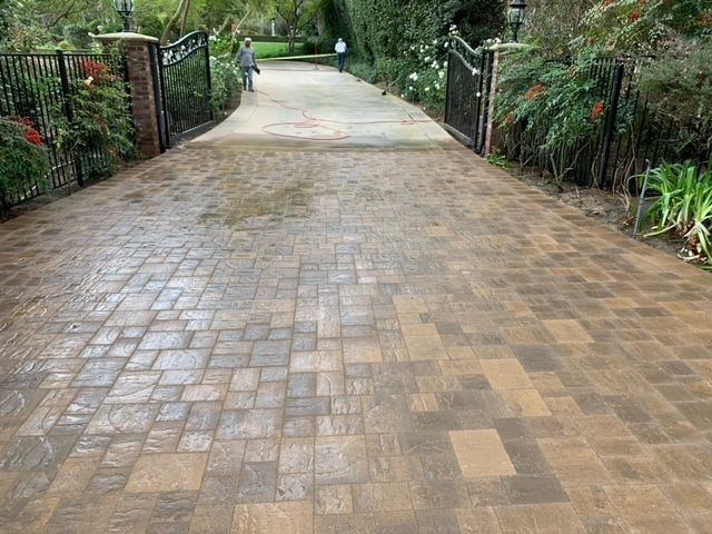 Paved driveway with open black gates, flanked by greenery and brick pillars; person walking in the distance.