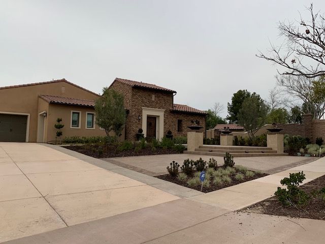 Tan stucco house with red-tile roof, stone facade, and paved driveway. Overcast sky.