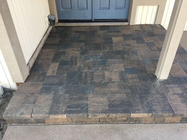 Brick paver porch with steps leading to a blue door. Beige pillars and siding surround the entrance.