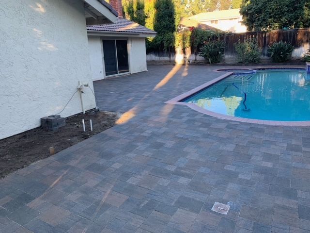 Poolside patio with dark paving stones. House, pool, and plants in the background.