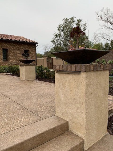 Concrete patio with planters atop stucco pillars, a brick building, and foliage in the background.