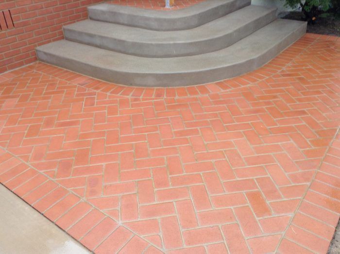 Red brick patio with curved concrete steps leading to a building. Herringbone pattern on the brick.