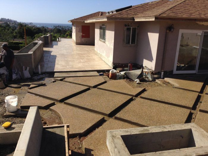 Construction site with concrete patio pavers being installed near a house with a brown roof and a view of the ocean.