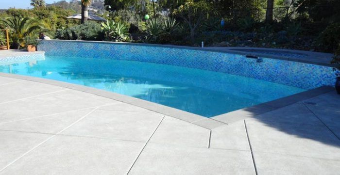 Pool with blue mosaic tile and light gray concrete decking under bright sunlight.