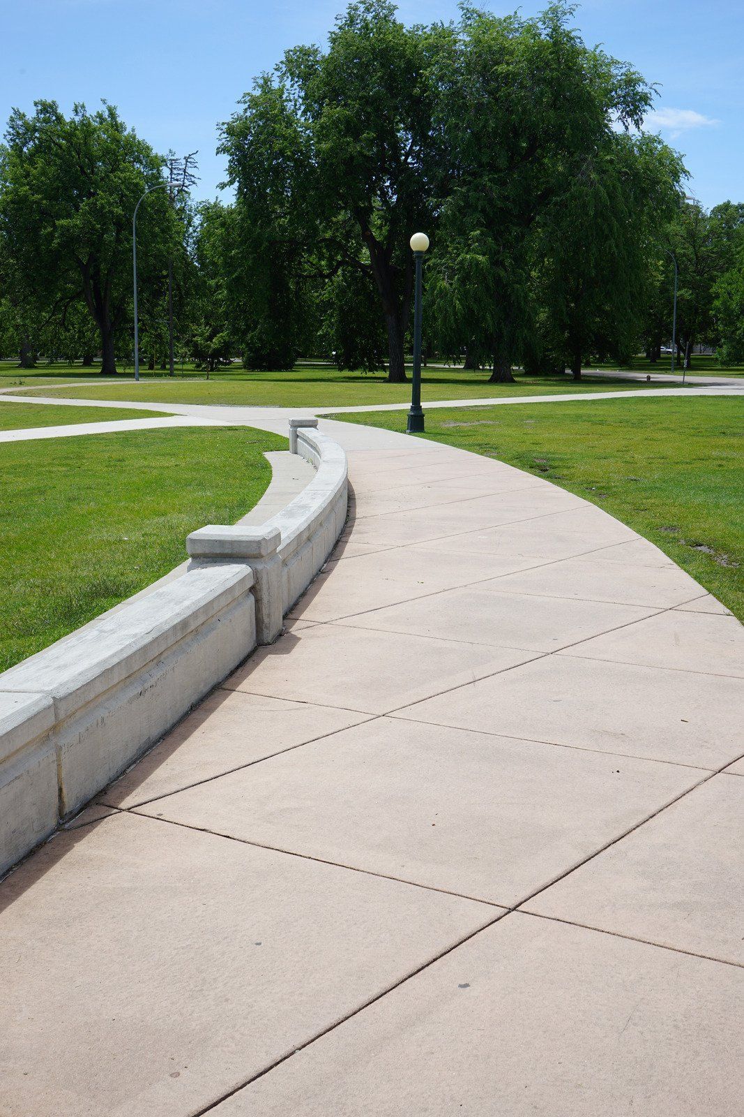 Curving concrete walkway with a low wall, leading through a grassy park. Trees and blue sky in background.