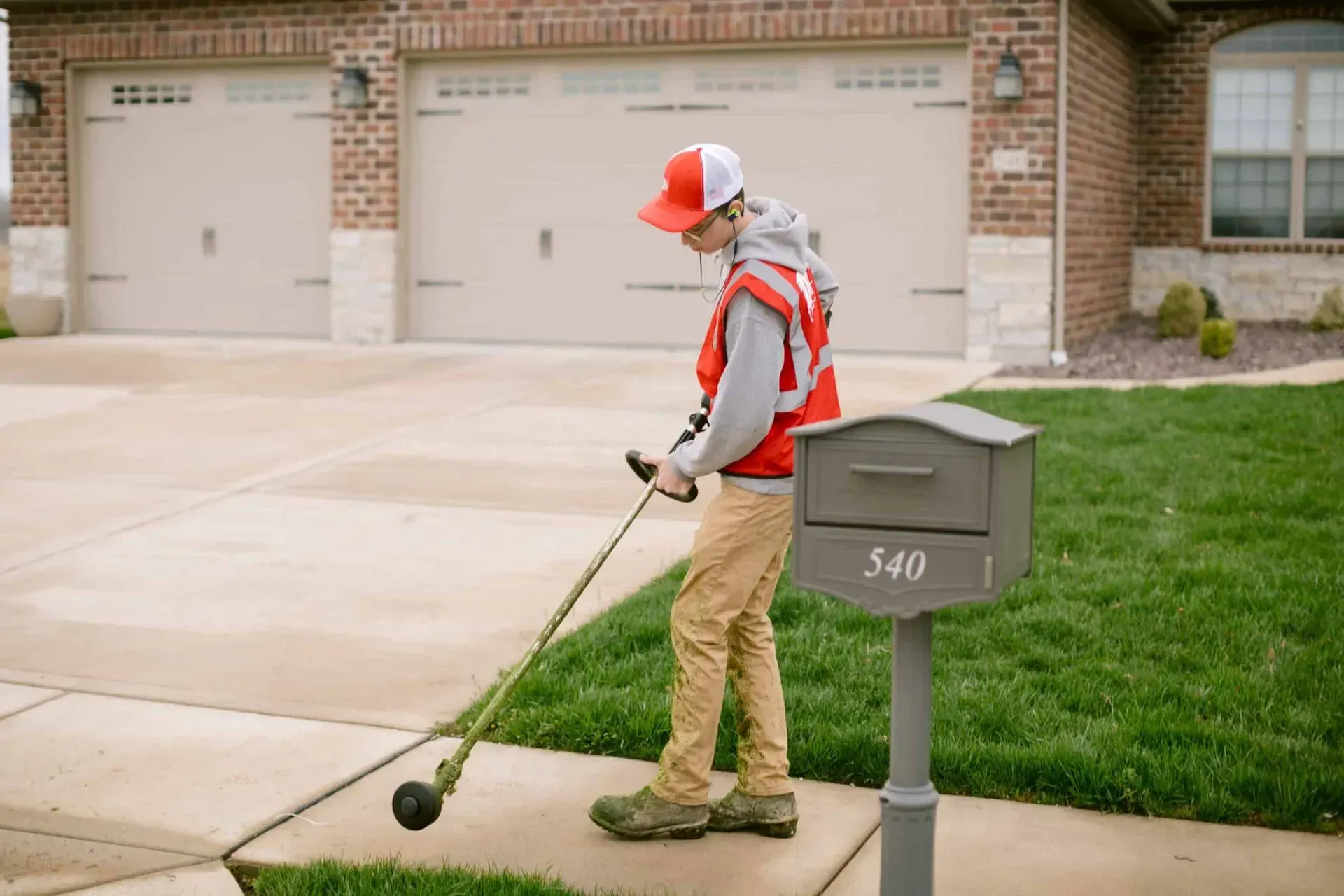 Young person in red safety vest using a weed eater along a sidewalk in front of a house.