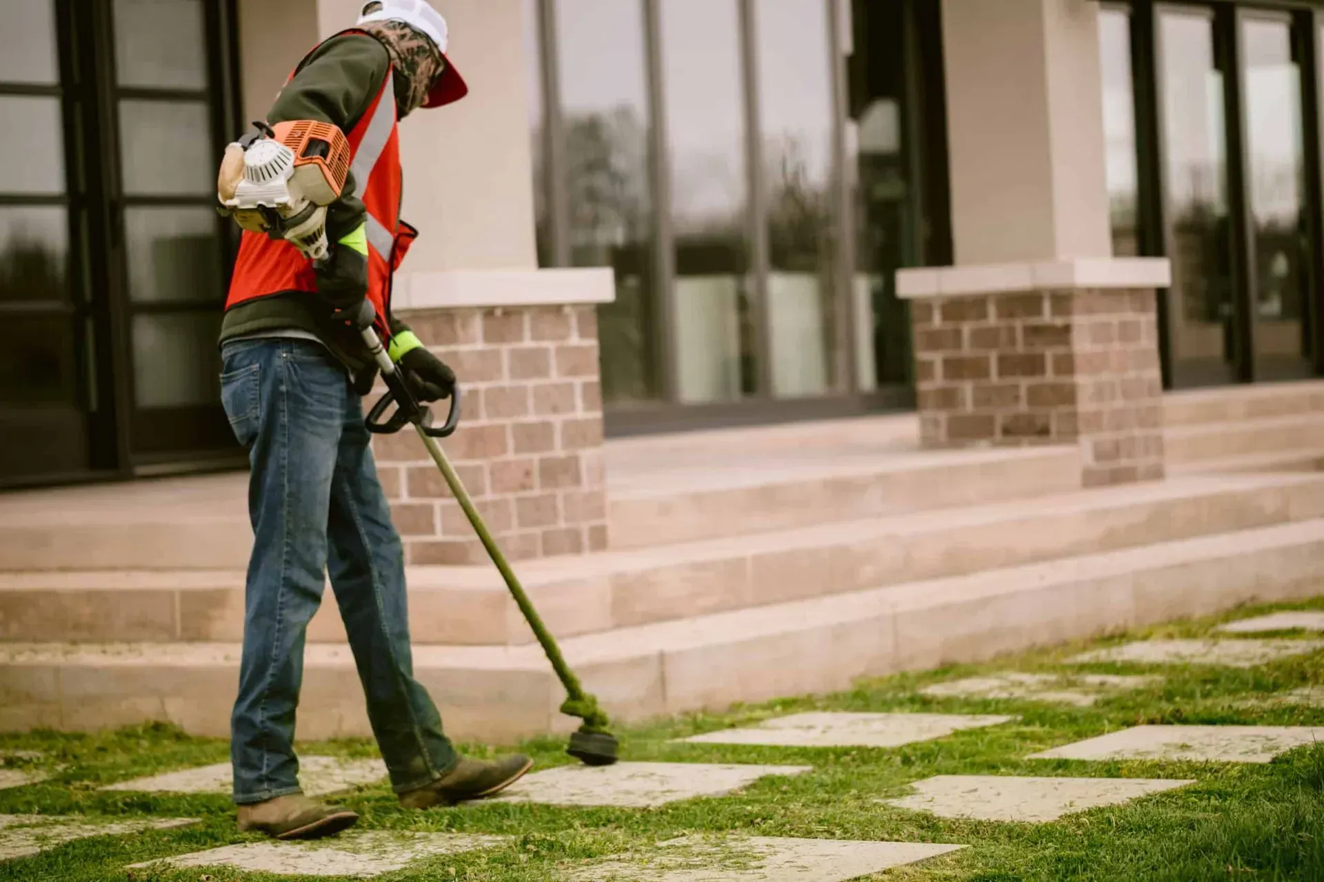 Person using a string trimmer to cut grass around stone pavers in front of a modern house.