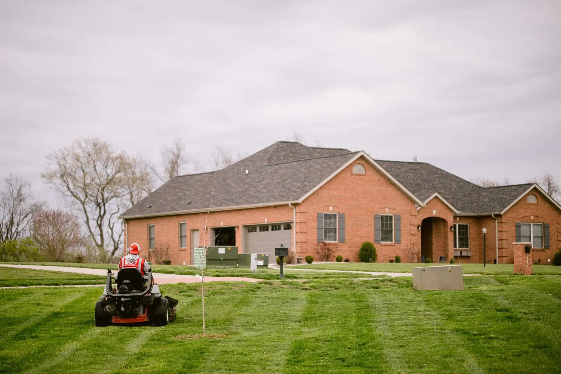 Person mowing lawn in front of a brick house with a gray roof on an overcast day.