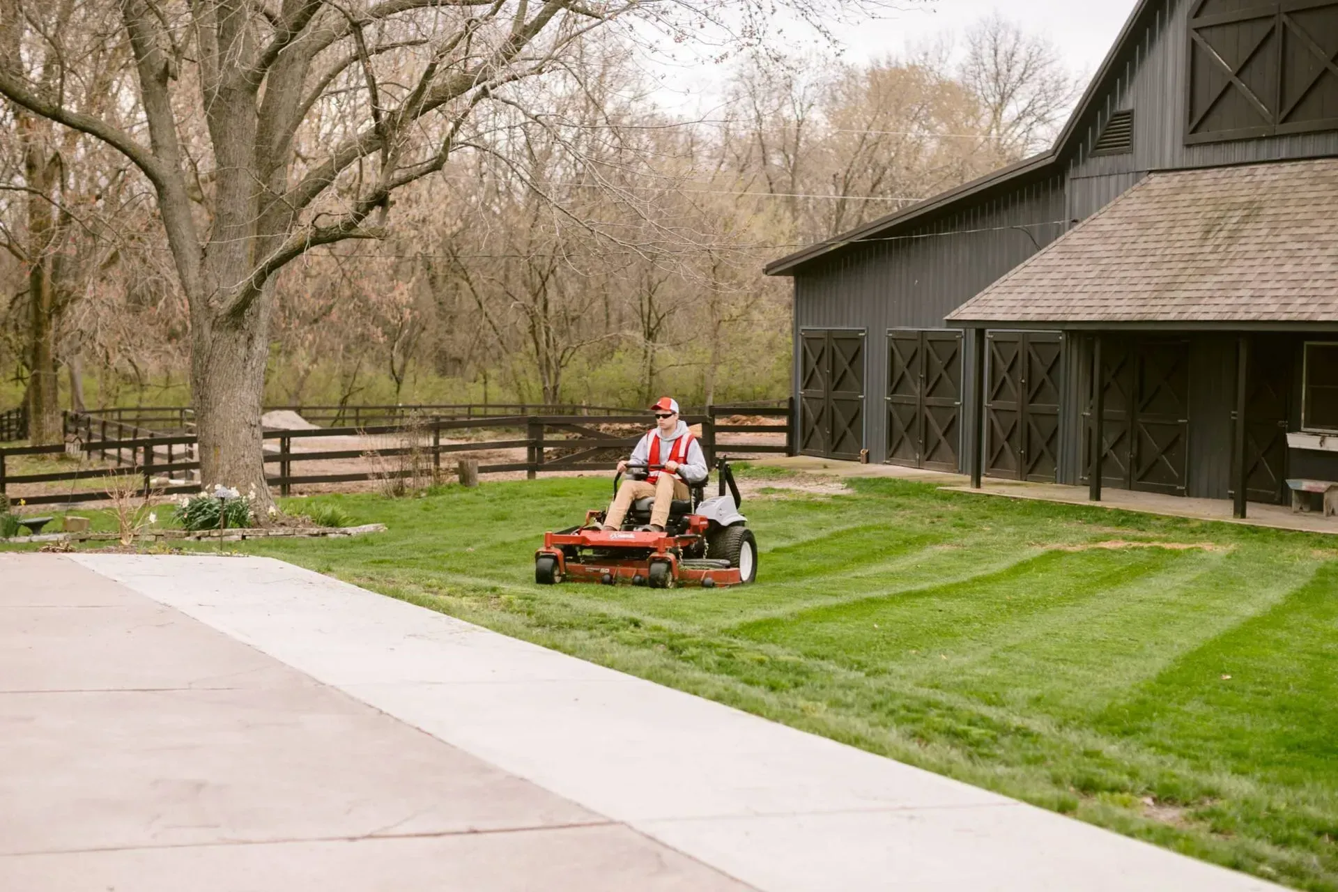 Man on a red riding lawnmower cuts the grass near a large barn in a sunny outdoor setting.