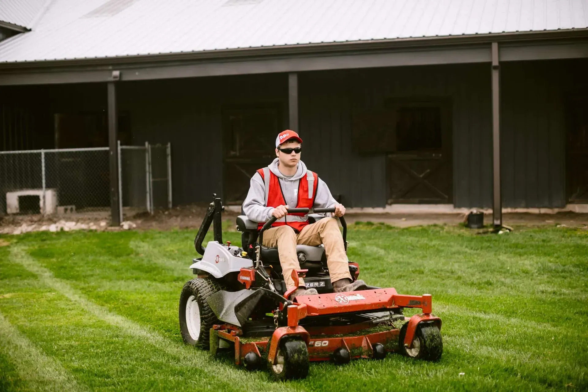 A person mowing grass on a riding lawn mower near a barn. They wear a safety vest and hat.