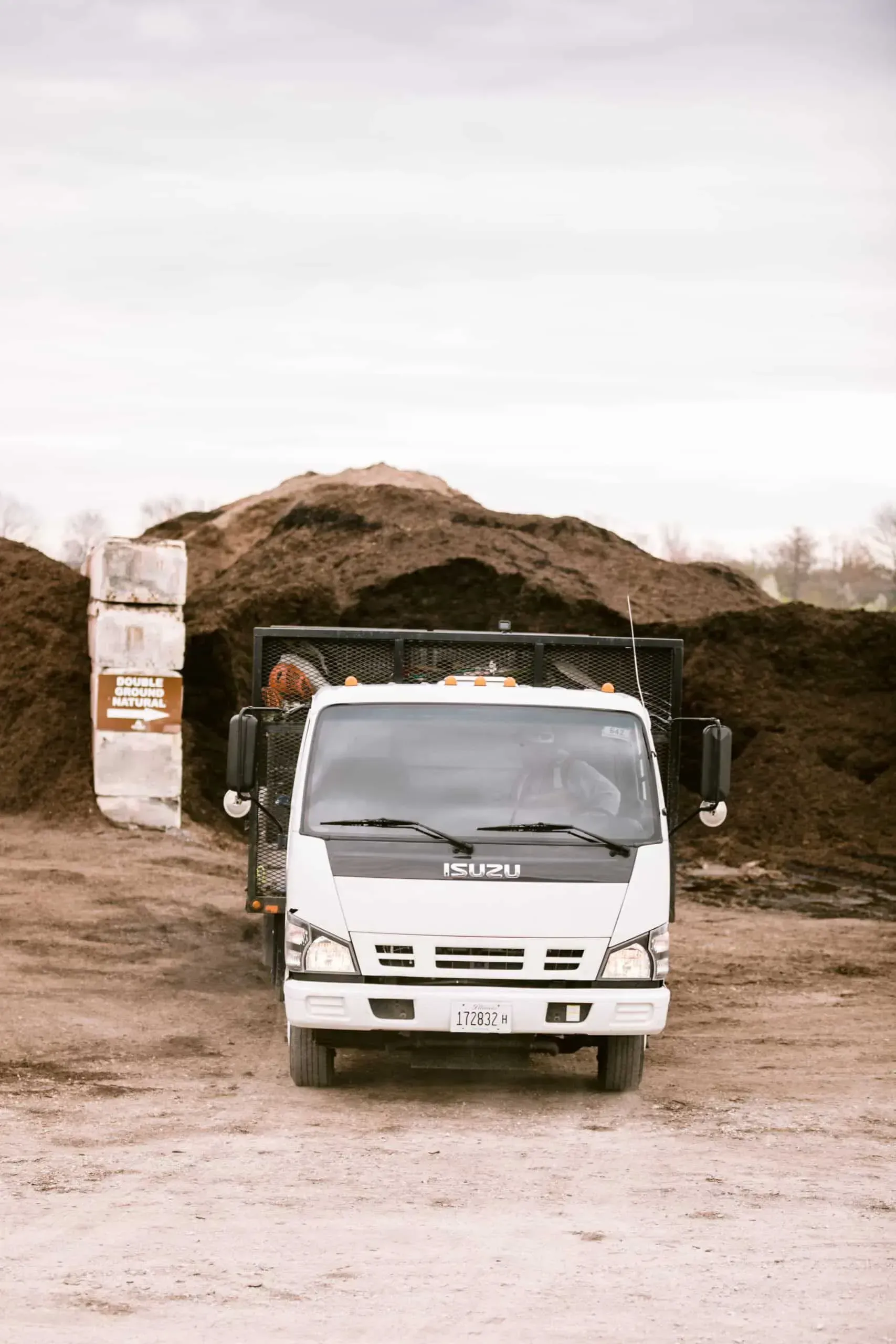 White Isuzu truck parked in front of piles of brown mulch on a cloudy day.