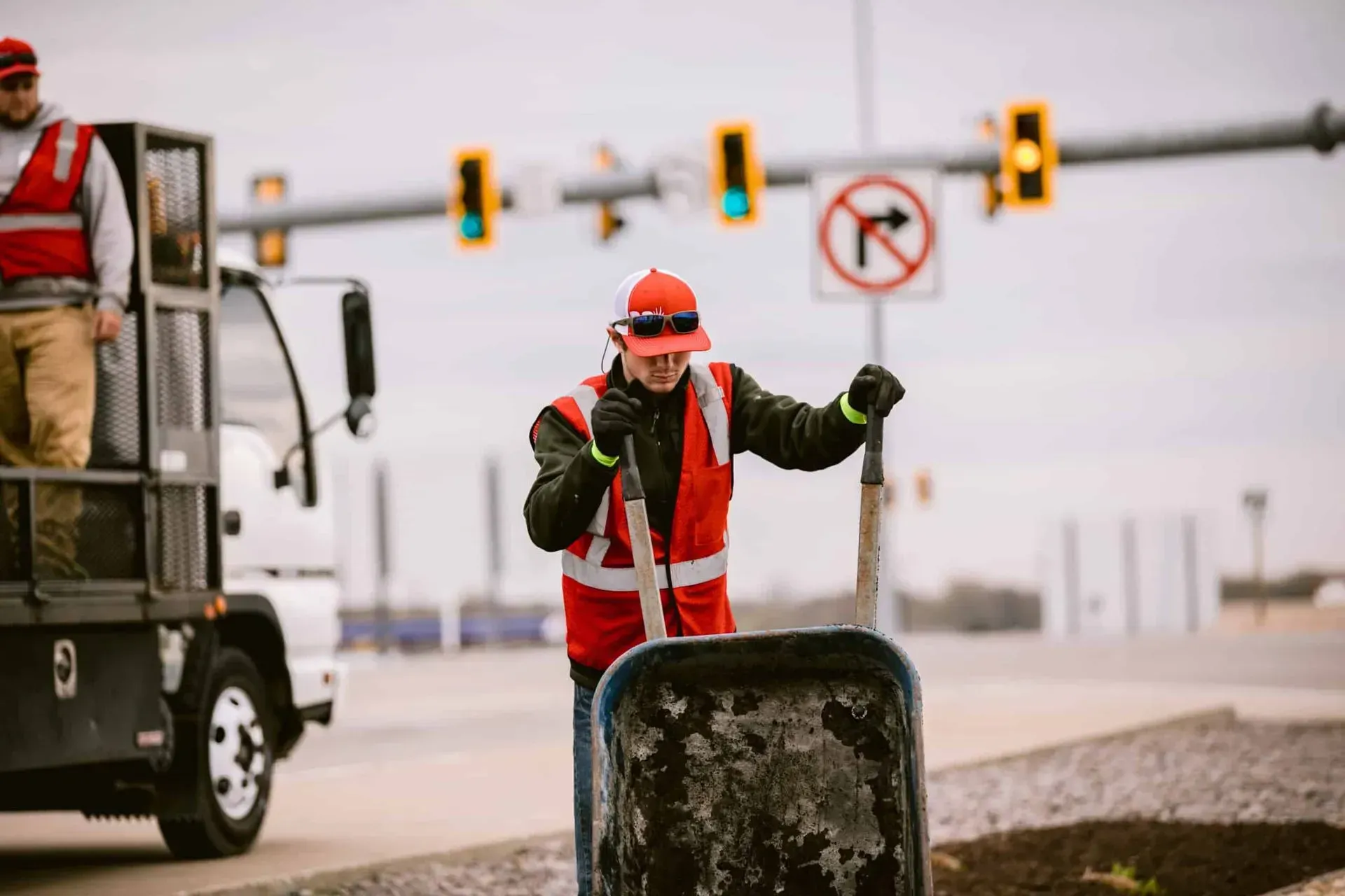 Man in red safety vest digging by a truck on the roadside, with traffic signals in the background.