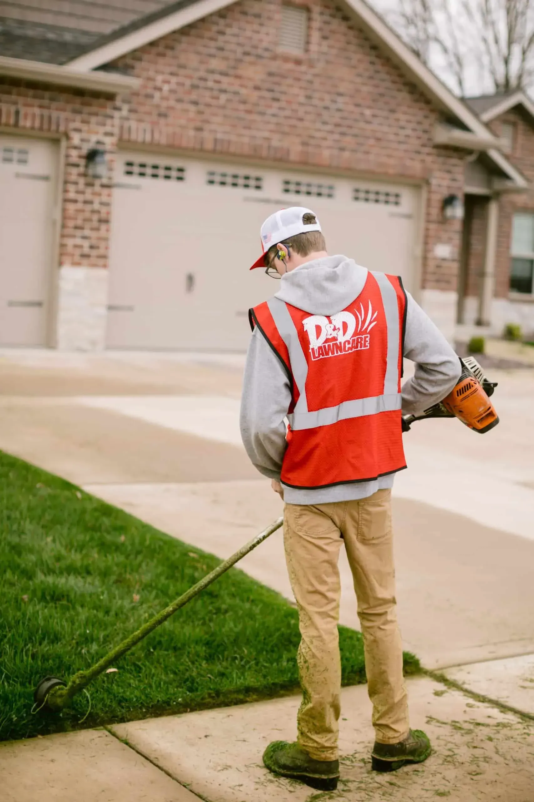 Landscaper using a weed whacker on a lawn; red vest, khaki pants, suburban home in background.