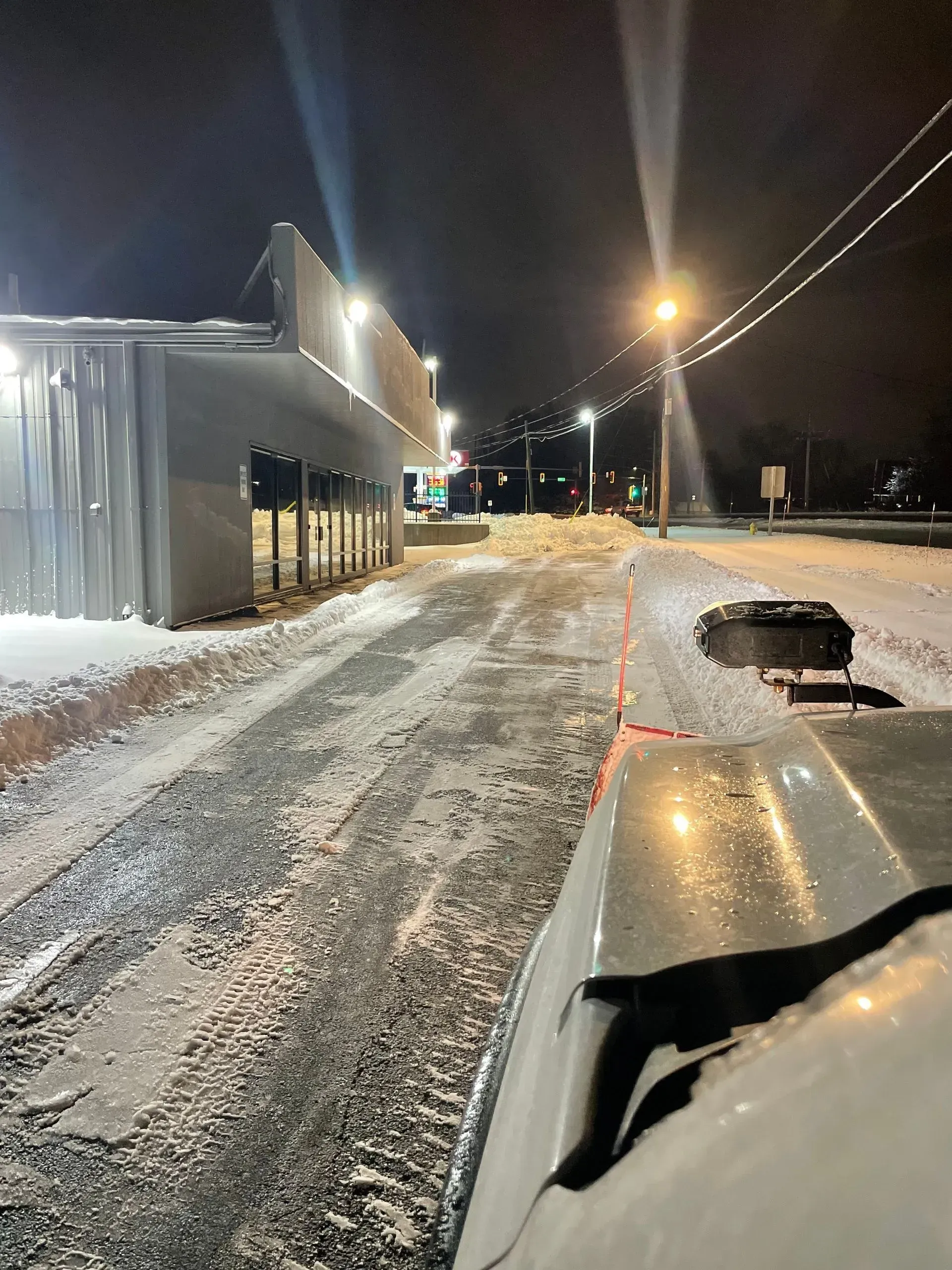 Snow-covered street at night with a building on the left, car dashboard in the foreground.