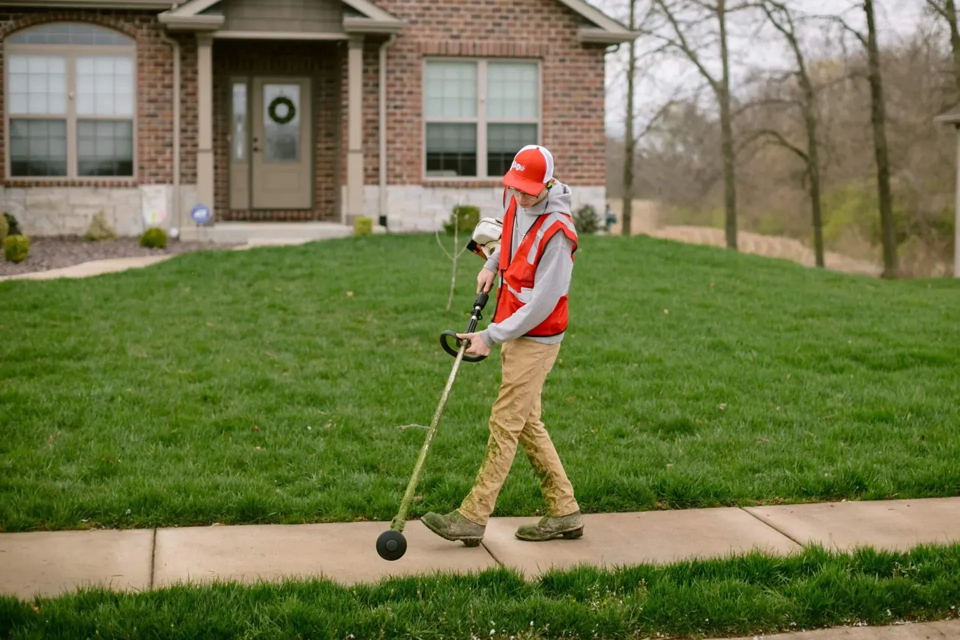 Person in safety gear using weed trimmer near house and sidewalk.
