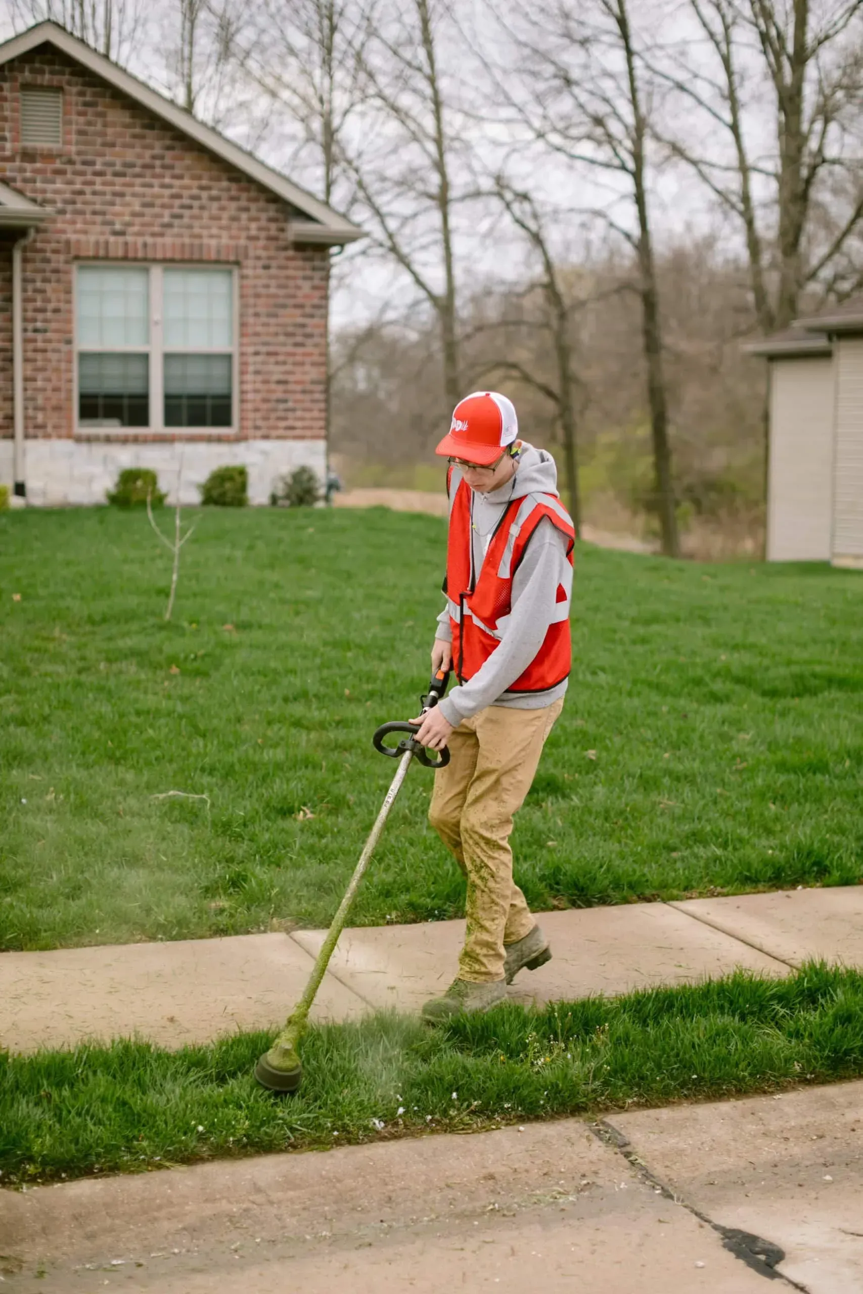 Man in orange safety vest trimming grass along a sidewalk. Brick house and lawn in the background.