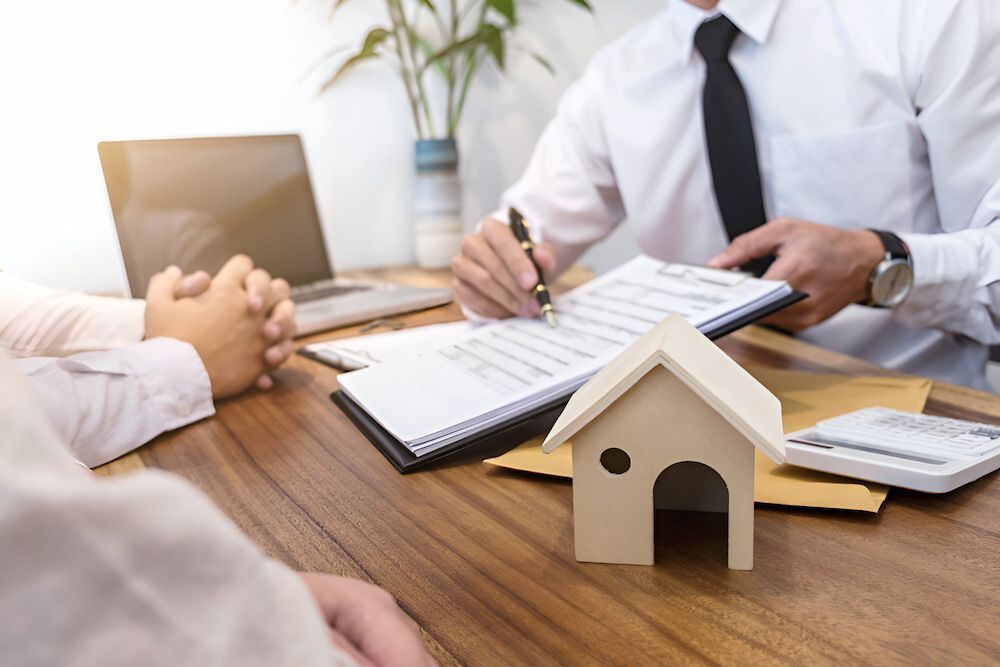 A Man Is Sitting At A Table Signing A Document Next To A Model House — Romy Sidhu Ray White Weir Views In Melton, VIC