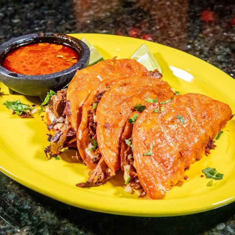 Three birria tacos with dipping sauce and lime wedges on a yellow plate.