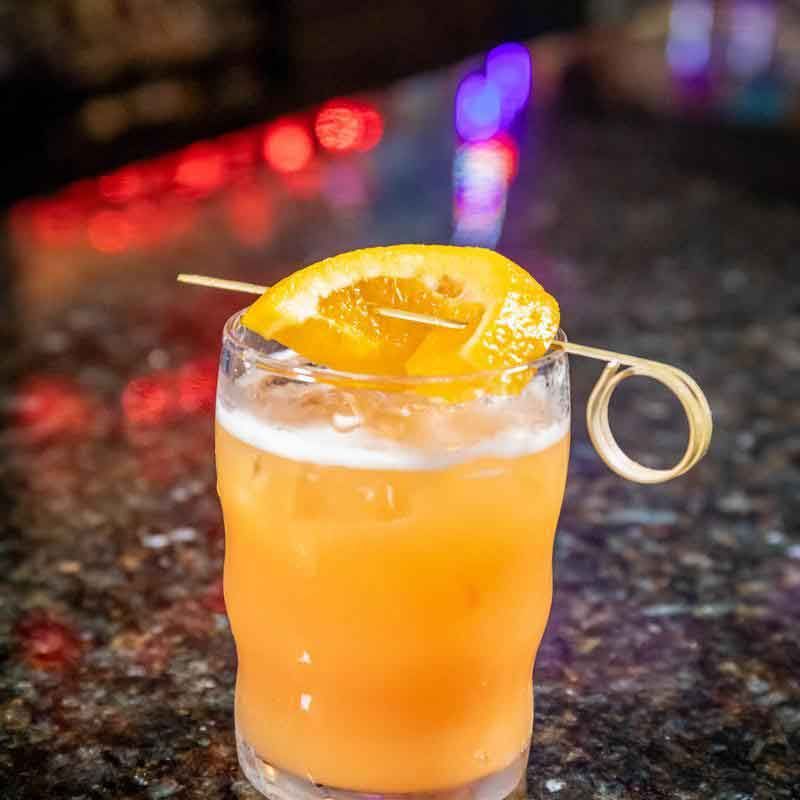 Cocktail in a glass garnished with an orange slice, set on a bar with blurred lights.