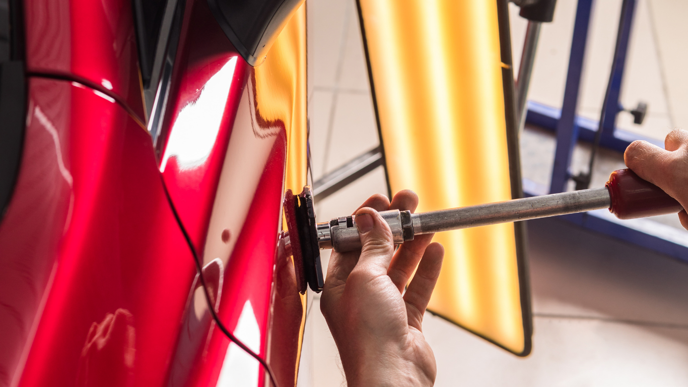 Person using a tool to repair a dent in a red car door, with a light in the background.
