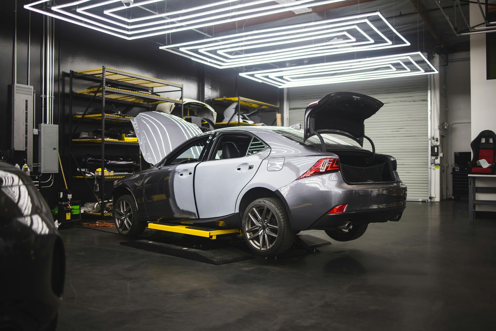A grey car with its hood and trunk open is raised on a lift inside a professional auto body shop under bright lighting.