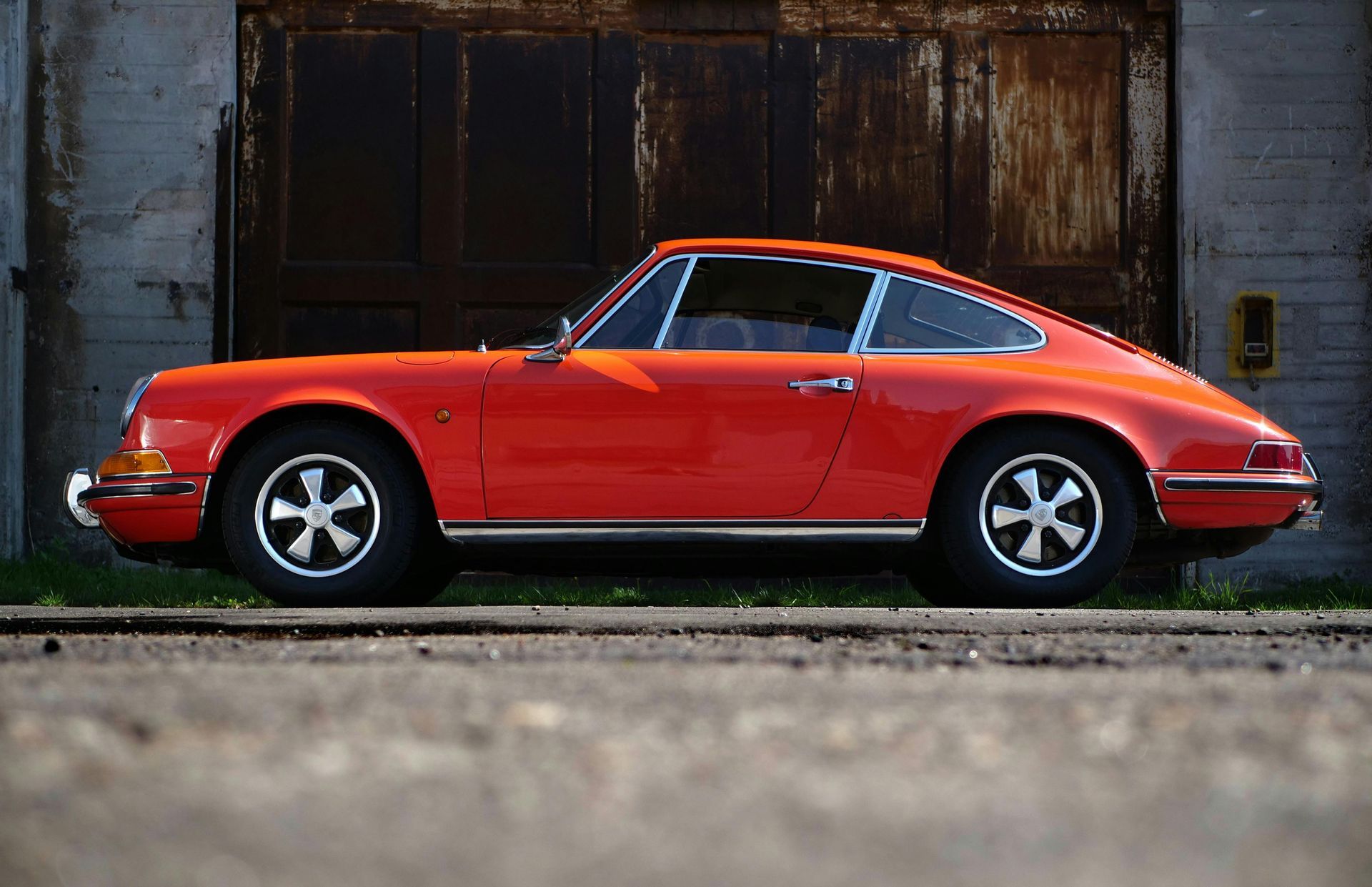 Red vintage Porsche 911 coupe parked in front of a weathered, dark garage door.