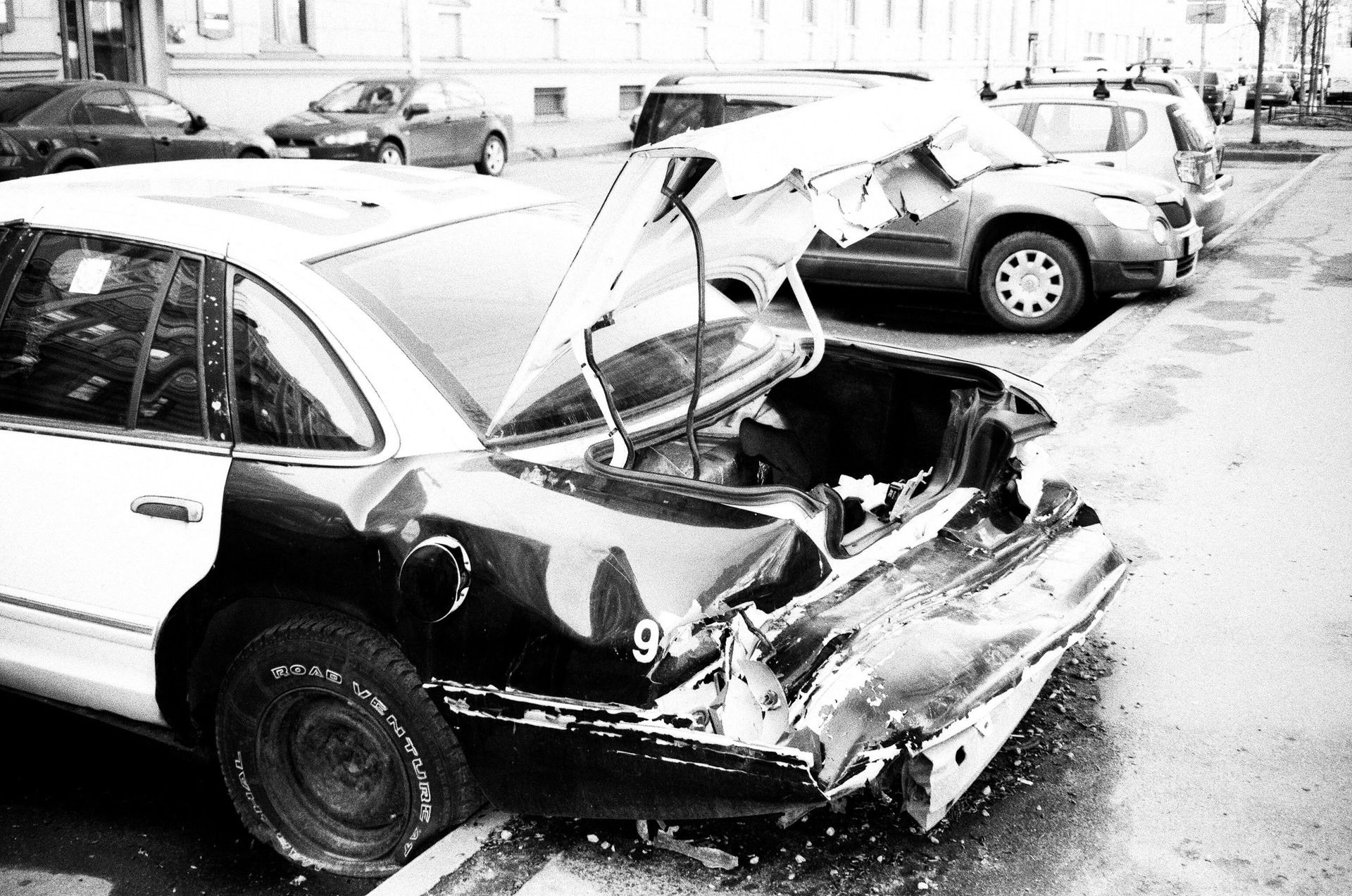 Damaged black and white police car, rear-end collision, trunk open, parked on a city street.