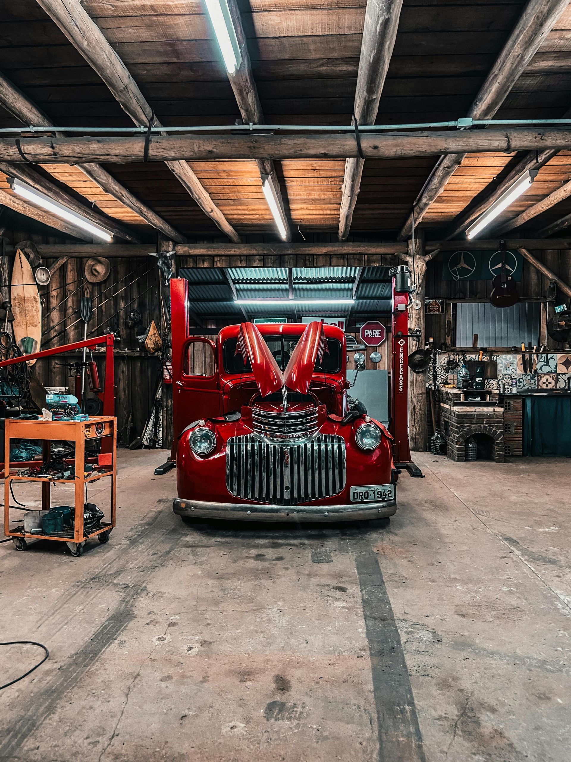 A bright red vintage truck with its hood open, parked in a rustic garage workshop with wooden beams.