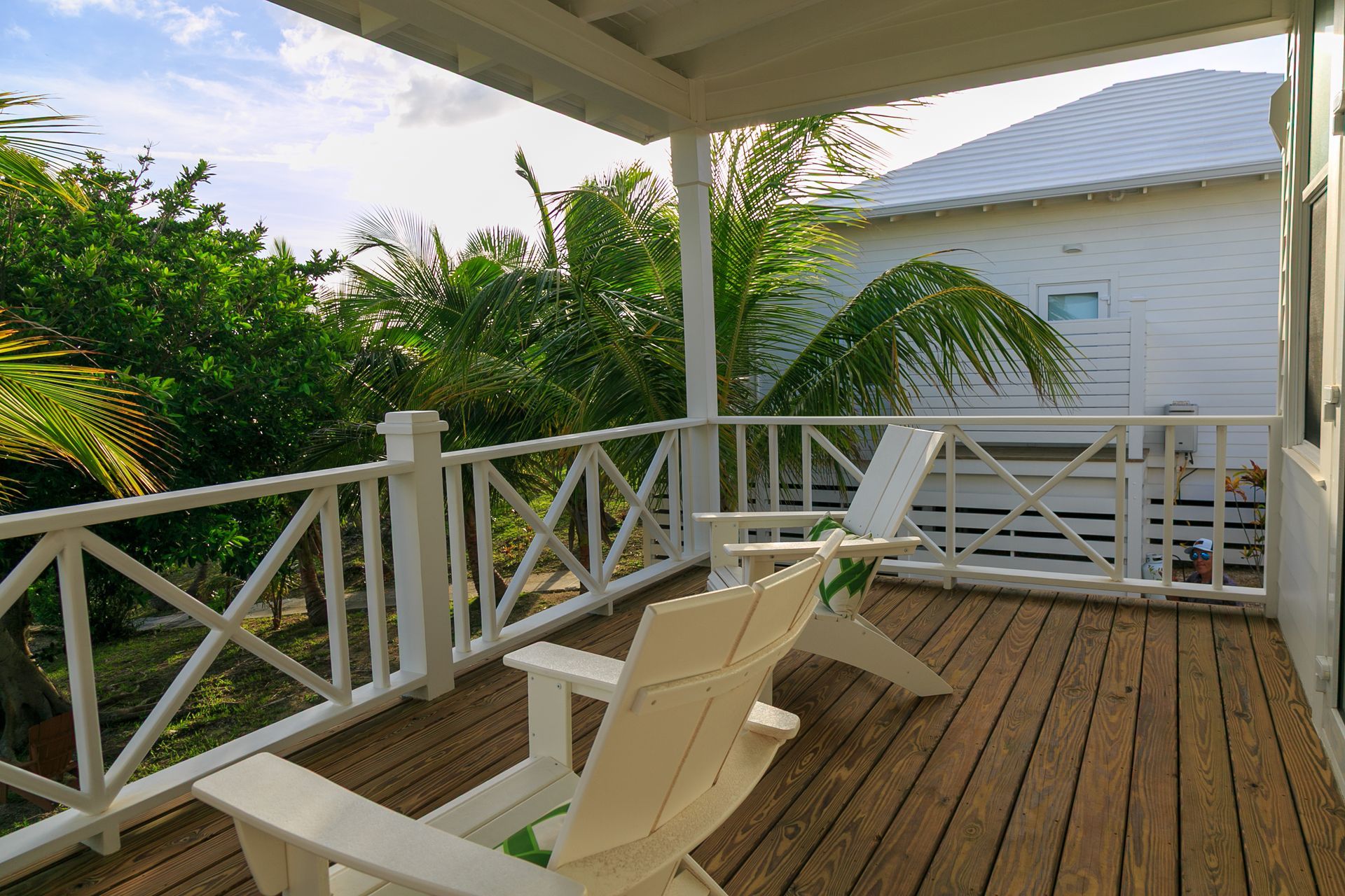 White Adirondack chairs on a wooden porch, overlooking lush green foliage and a white house.