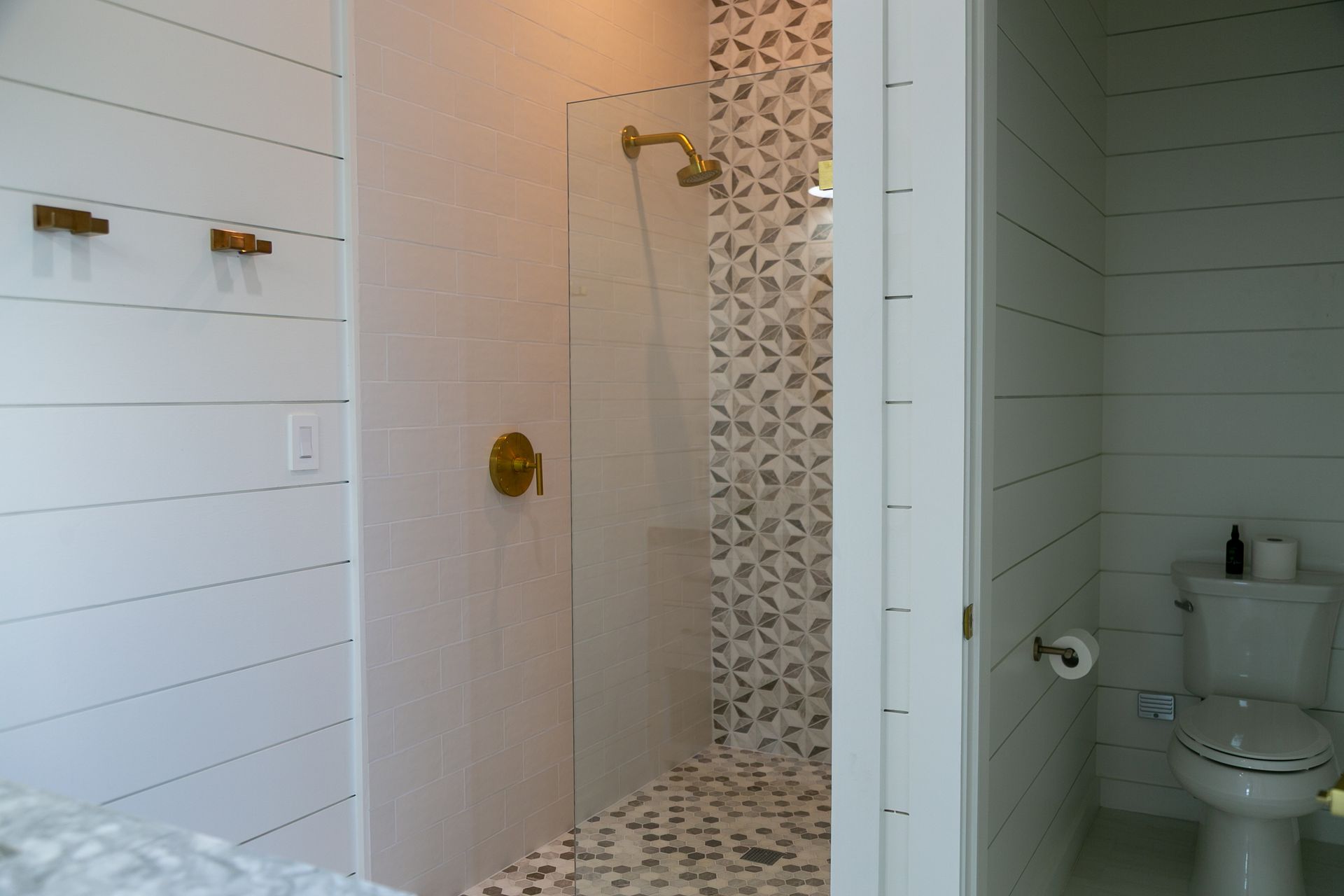 White-tiled bathroom with a glass shower, gold fixtures, pebble floor, and white shiplap walls. Toilet in the doorway.