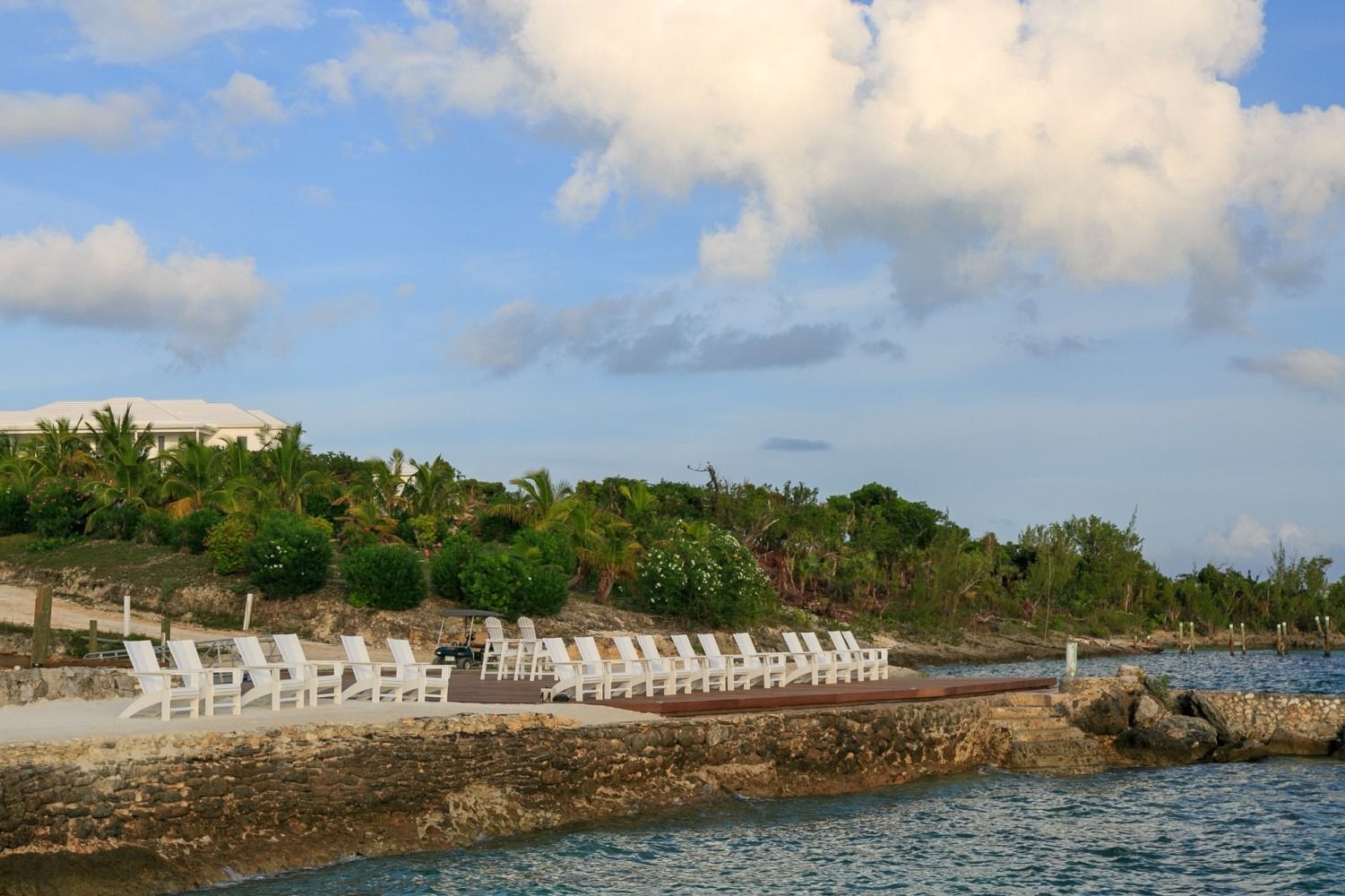 White lounge chairs on a stone pier, overlooking a calm blue ocean, under a partly cloudy sky.