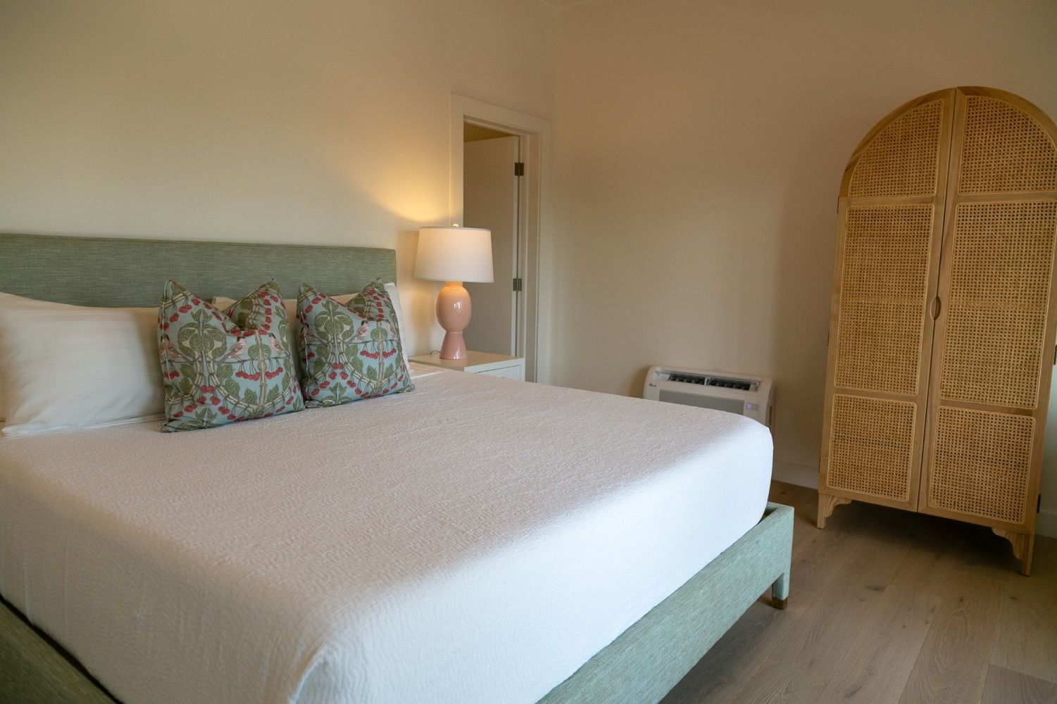 Bedroom with white bed, green headboard, patterned pillows, pink lamp, and woven wicker armoire.