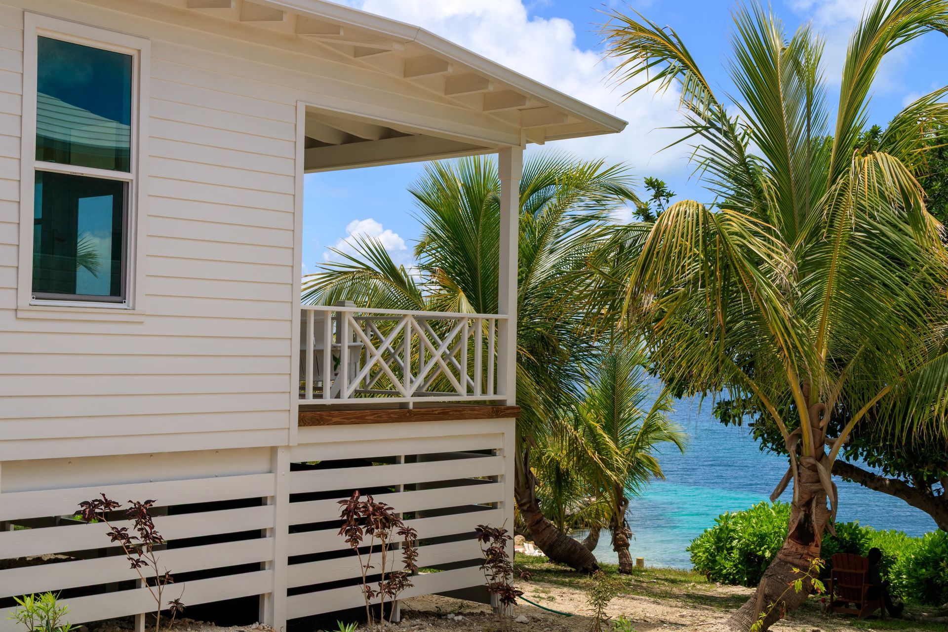 White cottage with balcony near a tropical beach; palm trees, blue water, and sky in background.