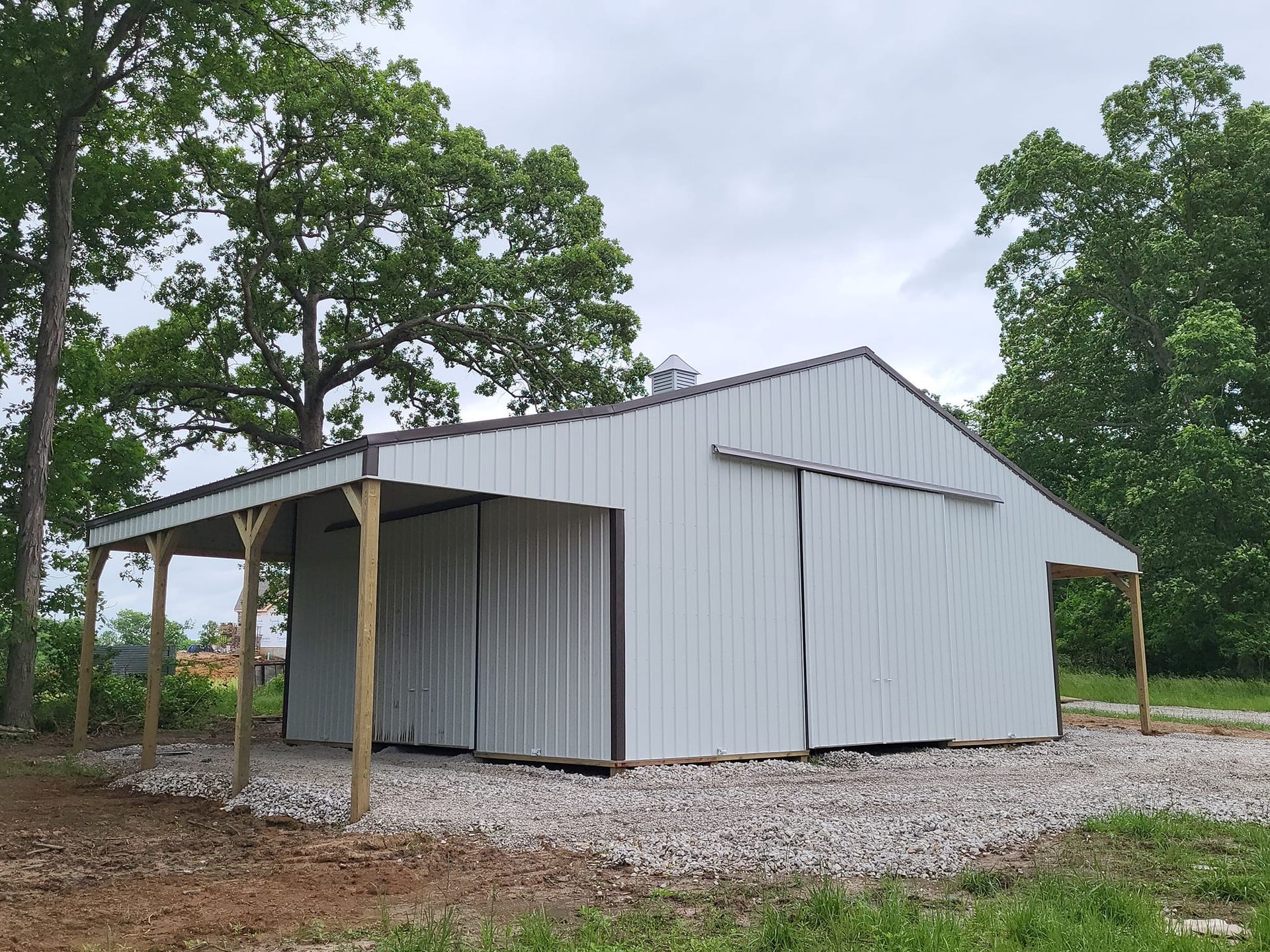 A white barn with a sliding door is surrounded by trees and gravel.